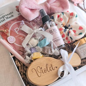 May include: A gift box containing a pomegranate facial mask, a pink plush headband, a clear spray bottle labelled Rose Water Toner, a white scrunchie with red and green heart patterns, a bag of wrapped sweets, and a wooden hairbrush engraved with Violet.