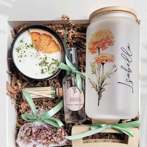 May include: A gift box containing a candle with dried orange slices, a small glass bottle of matches, a rose water spray bottle, a frosted glass tumbler with a bamboo lid featuring floral art and the name Isabella, and a beige hair claw clip.