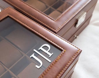 May include: Two brown rectangular watch storage boxes with clear glass lids and visible stitching. Each box features a grid of interior compartments with soft brown cushions. The glass lids are personalized with the silver-colored letters JIP.