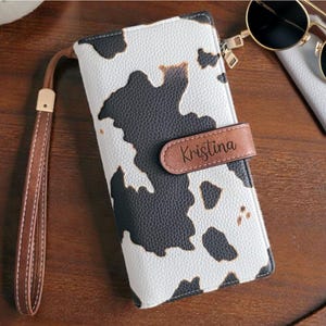 May include: A trifold PU leather wallet with a black and white cow print pattern. The wallet features a brown strap with the name Kristina engraved on the closure, a detachable brown hand strap, and measures 7.8 by 4.1 inches with RFID blocking.