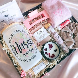 May include: A gift box containing a frosted glass tumbler with a bamboo lid and the text Nurse Justine, a pink hair claw clip, fuzzy pink socks, a lip balm, a beige scrunchie, a bar of soap, a vial of bath salts, a candle with rose petals, and matches.