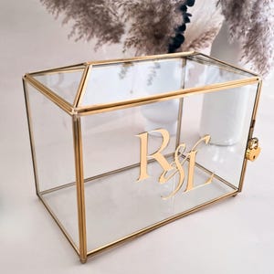 May include: A rectangular glass box with a gold metal frame and a hinged lid. The front panel features the gold-colored letters R&L in a script font. A small gold heart-shaped padlock is attached to the side of the frame to secure the box.
