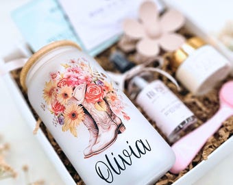 May include: A gift box containing a frosted glass tumbler with a bamboo lid, featuring a floral cowboy boot graphic and the name Olivia in black script. Also included are a small spray bottle, a cream jar, and a pink applicator tool.