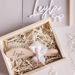 May include: A wooden hairbrush with soft bristles, personalized with the name Leyla and a small bow icon, tied with a pale pink ribbon. The brush rests in a wooden box filled with shredded paper, accompanied by a clear acrylic sign reading Leyla.