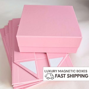 May include: A stack of flat-packed, light pink square boxes with one fully assembled box resting on top. The boxes feature magnetic closures and white adhesive strips visible on the folded flaps. The text LUXURY MAGNETIC BOXES FAST SHIPPING is present.