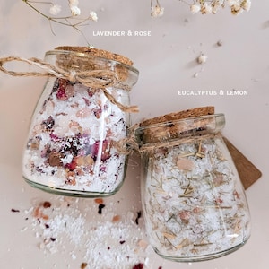 May include: Two glass jars filled with bath soak salts, one labeled "Lavender & Rose" and the other "Eucalyptus & Lemon." Each jar is tied with twine and has a cork lid. The text "3.4oz EACH" and "NATURAL BATH SOAK SALT SET" are also visible.