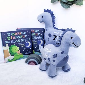 May include: Two blue plush dinosaur toys with dark blue spots and spikes, personalized with white text including the name Delilah Paige and birth details. Beside them are two copies of the book Dinosaur, Dinosaur, Say Good Night and a small rainbow.
