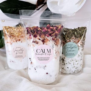 May include: Three clear stand-up pouches filled with bath soak. The left pouch is labeled Vanilla Peach, the center pouch is labeled CALM lavender & rose, and the right pouch is labeled Eucalyptus Lemon. Each contains salt and dried botanicals.