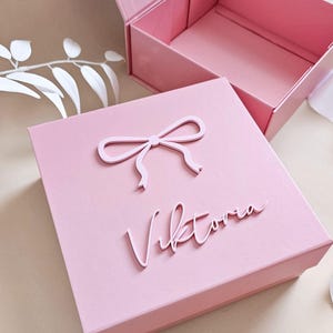 May include: A square pink gift box with a hinged lid, featuring a raised pink bow icon and the name Viktoria in raised script lettering on the top surface. The box is open, revealing a matching pink interior and a sturdy, structured design.