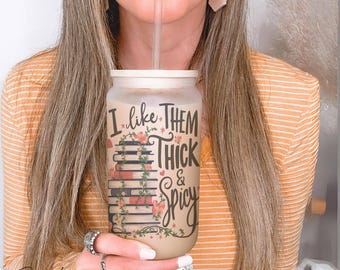 May include: A frosted glass tumbler with a bamboo lid and a clear straw, featuring a stack of books wrapped in floral vines and the black text I LIKE THEM THICK & SPICY printed on the front, filled with a light brown beverage.