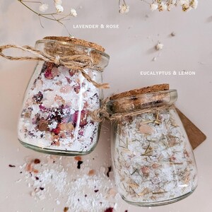 May include: Two glass jars filled with natural bath soak salt, each weighing 96g. One jar labelled Lavender & Rose contains white salt, pink crystals, and dried flower petals. The other labelled Eucalyptus & Lemon contains white salt, pink crystals, and dried herbs. Both jars feature cork stoppers and twine bows.