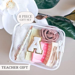 May include: A clear rectangular pouch containing an 8 piece gift set. Inside are a floral pen, a white fuzzy item, a dusty rose fabric flower, a beige hair claw clip, a pink rectangular object, and a white letter A patch with gold glitter trim.