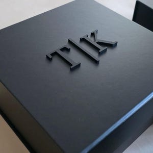 May include: A square black gift box with a matte finish featuring the raised, three-dimensional black letters T, I, and K centered on the lid. The box has a clean, structured design with sharp edges and a smooth, uniform surface texture throughout.