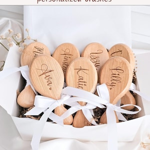 May include: A collection of light-colored wooden hairbrushes with oval heads and short handles, each engraved with a unique name in cursive script. The brushes are arranged in a white box and tied with white satin ribbons.