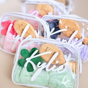 May include: Clear zippered pouches containing a wooden hairbrush, a plush headband, and a flower-shaped hair clip. Each pouch features a white name decal on the front, including names like Kylie, Lyla, and Karlie, with white ribbon zipper pulls.