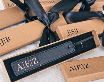 May include: A black rectangular leather bookmark with the embossed letters A|E|Z, presented in a brown cardboard gift box with a black satin ribbon. The bookmark features a matching leather strap and is displayed alongside other similar gift boxes.