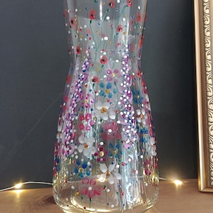 May include: A clear glass vase decorated with hand-painted floral patterns in red, pink, purple, blue, and white. The vase has a flared shape and is adorned with delicate flowers and dots. The vase is on a wooden surface.