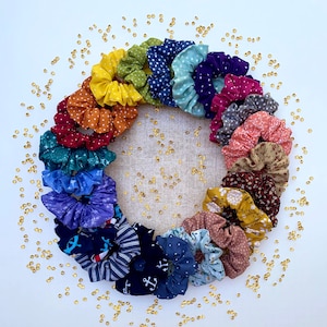 May include: A collection of 20 colorful fabric scrunchies in a variety of patterns, including polka dots, floral prints, and stripes. The scrunchies are arranged in a circle on a white background with gold glitter scattered around them.