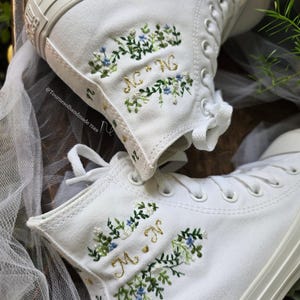 Peut inclure: Baskets montantes blanches avec broderie florale personnalisée. La broderie comprend des feuilles vertes, des fleurs bleues et des initiales dorées. Les baskets ont des lacets blancs et une semelle en caoutchouc blanche. Le texte "@Tournesolhandmade 7589" est visible.