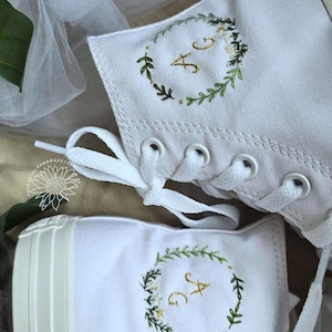 Peut inclure: Baskets montantes blanches avec des initiales brodées dans une couronne florale verte. Les baskets ont des lacets blancs et une semelle en caoutchouc blanche. Le fond comprend du tulle blanc et de la verdure.