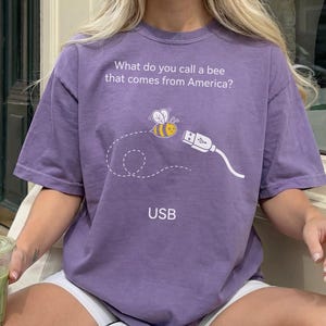 May include: A purple short-sleeve t-shirt featuring the white text 'What do you call a bee that comes from America?' above a graphic of a bee flying toward a USB cable connector, with the word 'USB' printed in white below the graphic.
