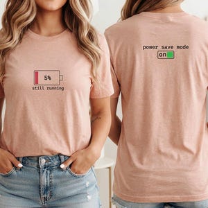 Pode incluir: Uma t-shirt de manga curta rosa claro com um gráfico de um ícone de bateria fraca a 5% e o texto still running na frente. A parte de trás da t-shirt apresenta o texto power save mode on com um ícone de bateria verde.
