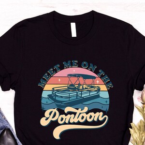 May include: Black t-shirt with a retro design featuring a pontoon boat on water, a sunset backdrop, and the text "Meet Me On The Pontoon." The shirt is paired with blue jeans and black shoes.