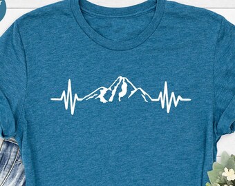 Könnte beinhalten: Ein T-Shirt in der Farbe Heidetiefblau mit einem weißen Bergketten-Motiv und einer Herzschlaglinie. Das Motiv ist eine stilisierte Bergkette mit einem Gipfel in der Mitte.