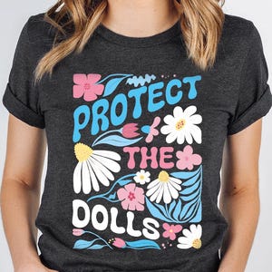 May include: Dark gray t-shirt with the text "PROTECT THE DOLLS" in a colorful floral design. The design includes pink, blue, and white flowers with green leaves. The shirt is short-sleeved and has rolled-up sleeves.