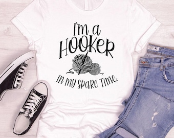 Op de afbeelding: Een wit T-shirt met korte mouwen en zwarte tekst I'm a HOOKER in my spare Time met een afbeelding van twee bollen wol en een haaknaald. Het T-shirt wordt getoond naast een blauwe spijkerbroek en zwart-witte sneakers.