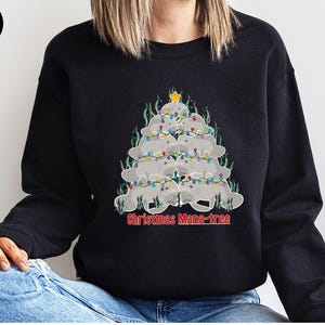 May include: A black crewneck sweatshirt featuring a graphic of grey manatees stacked in a triangular tree shape, decorated with colorful string lights and a yellow star on top. The text "Christmas Mana-tree" is printed in red below the graphic.