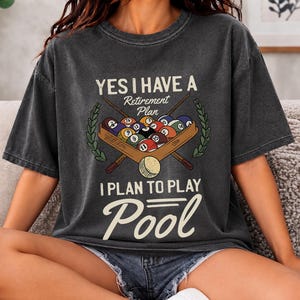 Peut inclure: Un t-shirt gris anthracite avec un motif de boules de billard et deux queues de billard croisées. Le texte indique : YES I HAVE A Retirement Plan I PLAN TO PLAY Pool. Le design comprend des branches de laurier vertes de chaque côté.