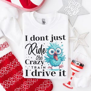 May include: White crew neck sweatshirt with the text "I don't just ride the crazy train, I drive it" and a cartoon blue owl. The image is surrounded by Christmas decorations, including a stocking and a Santa-themed wine glass.