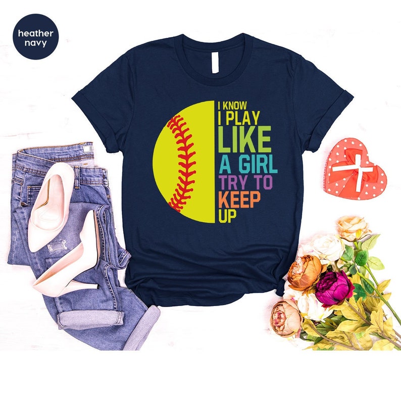 K&ouml;nnte beinhalten: Ein marineblaues T-Shirt mit einer gelben Softball-Grafik und dem Text "I know I play like a girl try to keep up". Der Text ist in Regenbogenfarben.