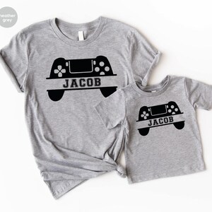 May include: Two heather grey t-shirts with a black video game controller graphic and the name "Jacob" printed on each shirt.