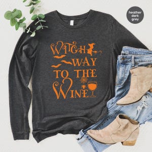 Op de afbeelding: Een donkergrijs gemêleerd shirt met lange mouwen en oranje tekst Witch Way To The Wine, vergezeld van grafische illustraties van een heksensilhouet, vleermuizen, een spinnenweb en een wijnglas, gecombineerd met een blauwe spijkerbroek en beige laarzen.