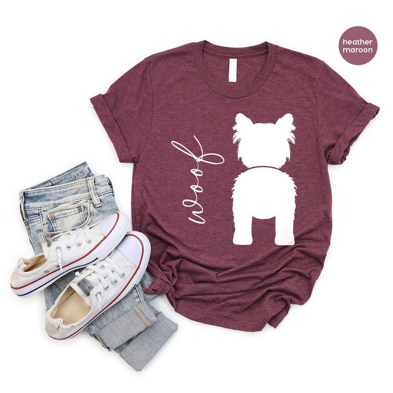 Puede incluir: Una camiseta granate con la silueta de un perro blanco y la palabra "Woof" en cursiva. La camiseta se combina con vaqueros azules y zapatillas blancas. La camiseta tiene mangas cortas y cuello redondo.
