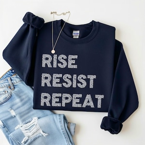 May include: Navy blue sweatshirt with the words "RISE RESIST REPEAT" in white, stylized text. The sweatshirt is folded on top of a pair of light blue jeans. A gold necklace with a pendant is draped over the sweatshirt.
