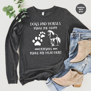 May include: A heather dark grey long-sleeve shirt featuring white text that reads DOGS AND HORSES MAKE ME HAPPY HUMANS MAKE MY HEAD HURT, accompanied by a white graphic of a dog paw print and a horse silhouette, paired with blue denim jeans.