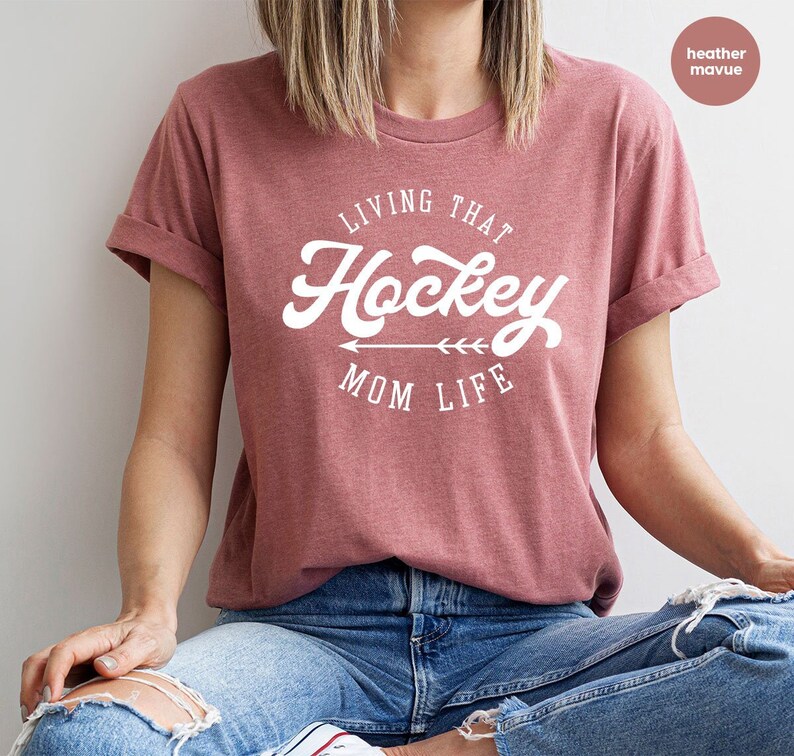 May include: A heather mauve t-shirt with the text "Living That Hockey Mom Life" in white lettering with a white arrow pointing to the right.