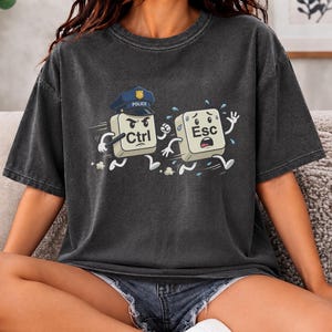 Peut inclure: Un t-shirt gris anthracite à manches courtes avec une impression graphique représentant une touche Ctrl de clavier déguisée en policier poursuivant une touche Esc. La touche Ctrl porte un képi bleu et tient une matraque, tandis que la touche Esc fuit.