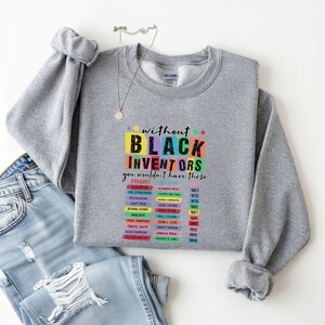 May include: A gray sweatshirt with the text "without BLACK INVENTORS you wouldn't have these" in colorful block letters. Below the text are lists of products and inventors. The sweatshirt is paired with blue jeans.