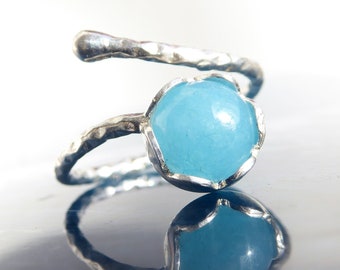 May include: A silver-toned ring with a light blue gemstone. The ring features a textured band that wraps around the finger and culminates in a bezel setting that holds the round, light blue stone. The ring is displayed on a reflective surface.