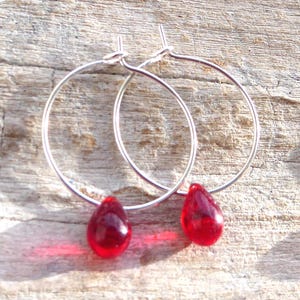 May include: Silver hoop earrings featuring teardrop-shaped, deep red glass beads. The earrings are set against a weathered, light brown wooden surface, contrasting the shiny metal with the rustic background.