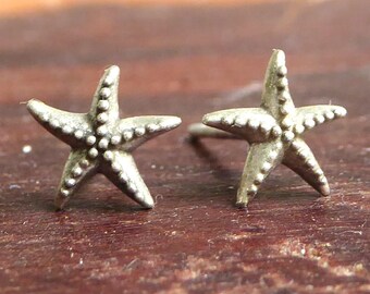 May include: A pair of silver-colored starfish stud earrings. Each earring features a detailed, textured design with small, raised dots. The earrings are set against a blurred wooden background.