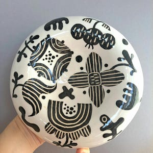 Op de afbeelding: Een ronde keramische kom met een glanzend witte glazuurlaag en handgeschilderde zwarte motieven. Het oppervlak toont diverse abstracte vormen, waaronder gestreepte bloemmotieven, geometrische figuren en verspreide stippen.
