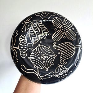 Può includere: Un piatto rotondo in ceramica nera caratterizzato da un disegno a linee bianche disegnato a mano di vari fiori stilizzati e forme botaniche. La superficie lucida mostra motivi complessi tra cui zigzag, punti e strisce nei motivi floreali.