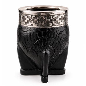 May include: A black mate gourd featuring a carved floral pattern on the body and a silver-toned metal rim decorated with a geometric cross design. The vessel is supported by three small feet at the base, providing a stable structure for this cup.