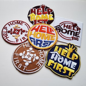 Op de afbeelding: Een verzameling geborduurde patches met de tekst "Help Home First" in verschillende kleuren en ontwerpen. De patches zijn rond en rechthoekig en bevatten afbeeldingen van huizen en handen. Het kleurenpalet omvat rood, geel, blauw en bruin.