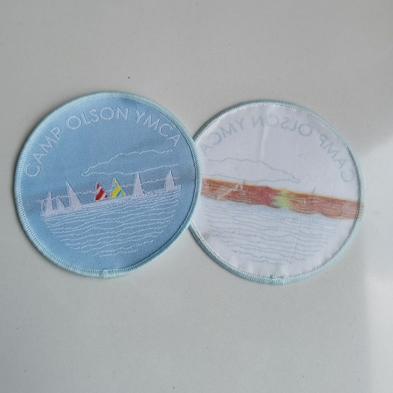 May include: Two round blue and white embroidered patches. One patch says "Camp Olson YMCA" and depicts sailboats on a lake. The other patch has a similar design but says "Camp Olson YMCA" in reverse.